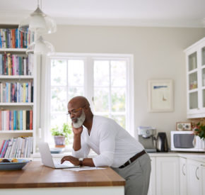 Older person using a laptop at counter in home with books and kitchen appliances in the background