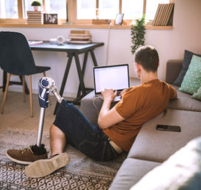 User with prosthetic left leg using laptop seated on the floor of a house on a sunny day