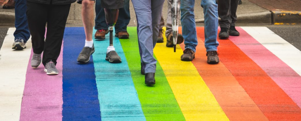 Eight people beginning to cross a rainbow flag painted street all depicted with legs only from the hip down and one person with a prosthetic left leg