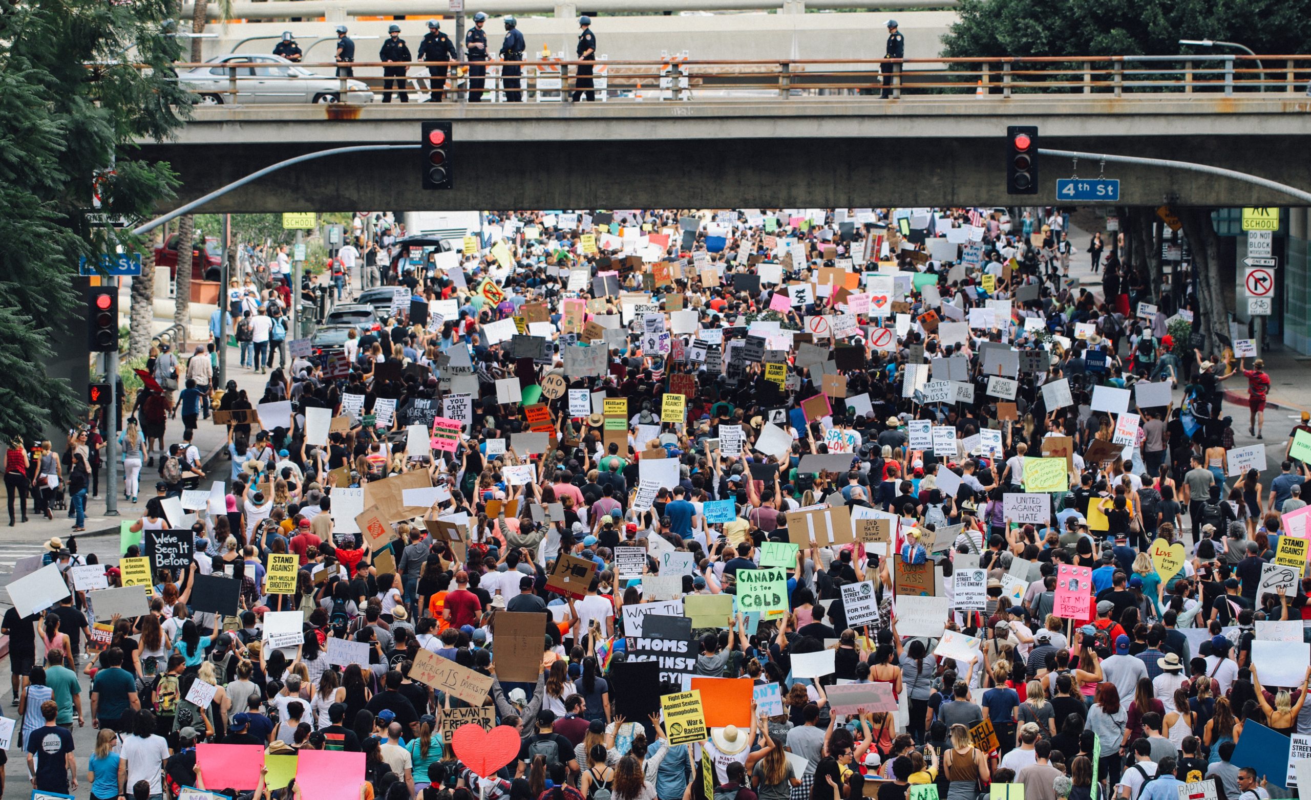 mass of people holding signs in a protest march walking under a bridge with police above on the bridge