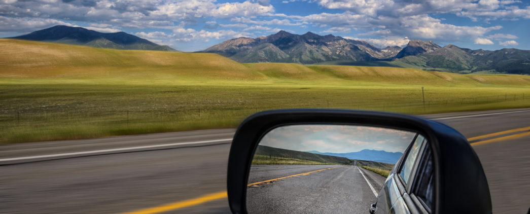 Side mirror of a vehicle with clear view of the road behind as the road ahead while larger view outside of mirror of grassy hills and mountains speed past in the distance