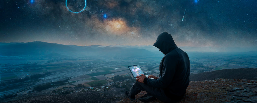 Hooded man with laptop on a rocky outcropping with night sky filled with galaxy of bright stars and planets
