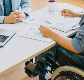 Man using wheelchair meets with someone behind a desk in an office environment.