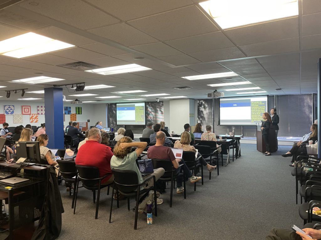 A group of people attend a class on document accessibility. There is a presentation shown on two screens  as an ASL interpreter translates for the instructor.