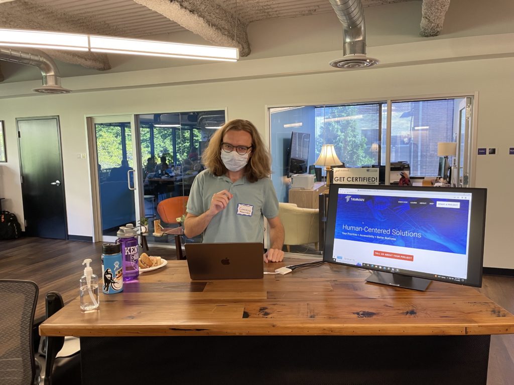 Tamman's Cameron Messinides stands at a desk with a laptop and a monitor to display how bad headings can impact screen reader users.