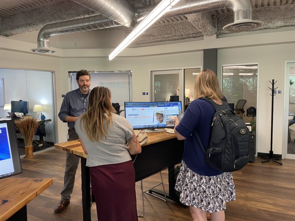 Mike Mangos delivers a demonstration of common web accessibility issues at a desk with a monitor as two onlookers listen. 