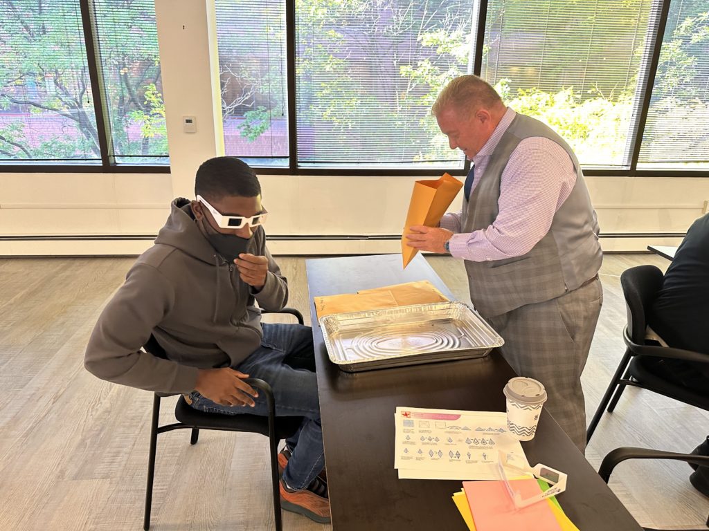 One man sits at a table wearing glasses that simulate a visual disability, across from him another man stands while looking into an envelope.