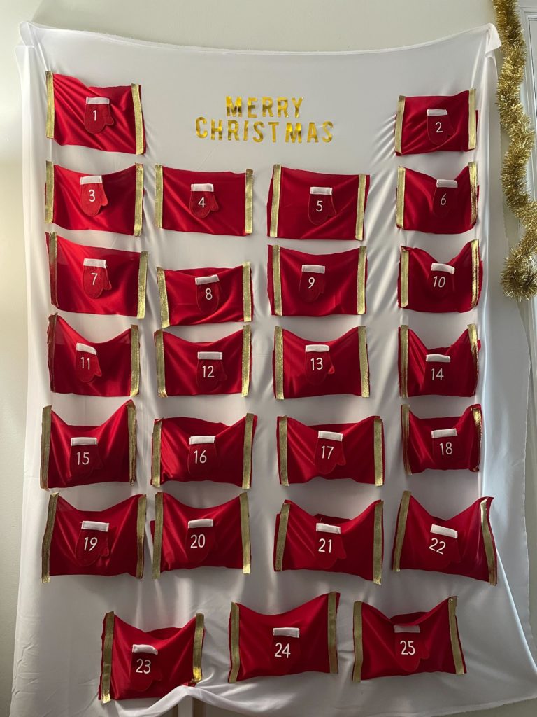 A large sheet with 25 red sock pouches, numbered 1-25 with a red mitten on them. At the top it says "Merry Christmas" in gold writing.