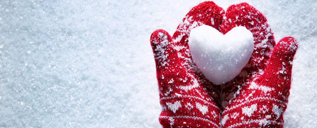 Red mittens holding a heart-shaped snowball on a snowy day.