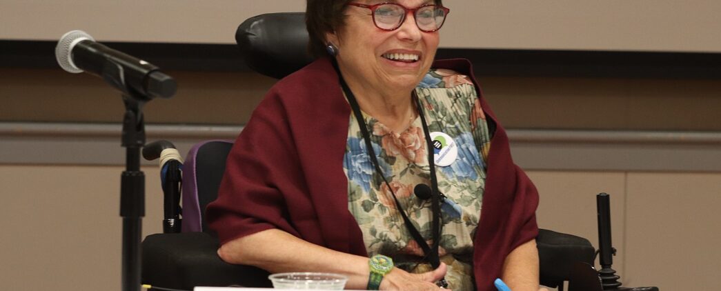 Disability advocate and pioneer Judith Heumann speaks at a conference. She is smiling, has red glasses on, is wearing a floral shirt, red shawl, and is sitting in her mechanical wheelchair.