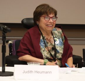 Disability advocate and pioneer Judith Heumann speaks at a conference. She is smiling, has red glasses on, is wearing a floral shirt, red shawl, and is sitting in her mechanical wheelchair.