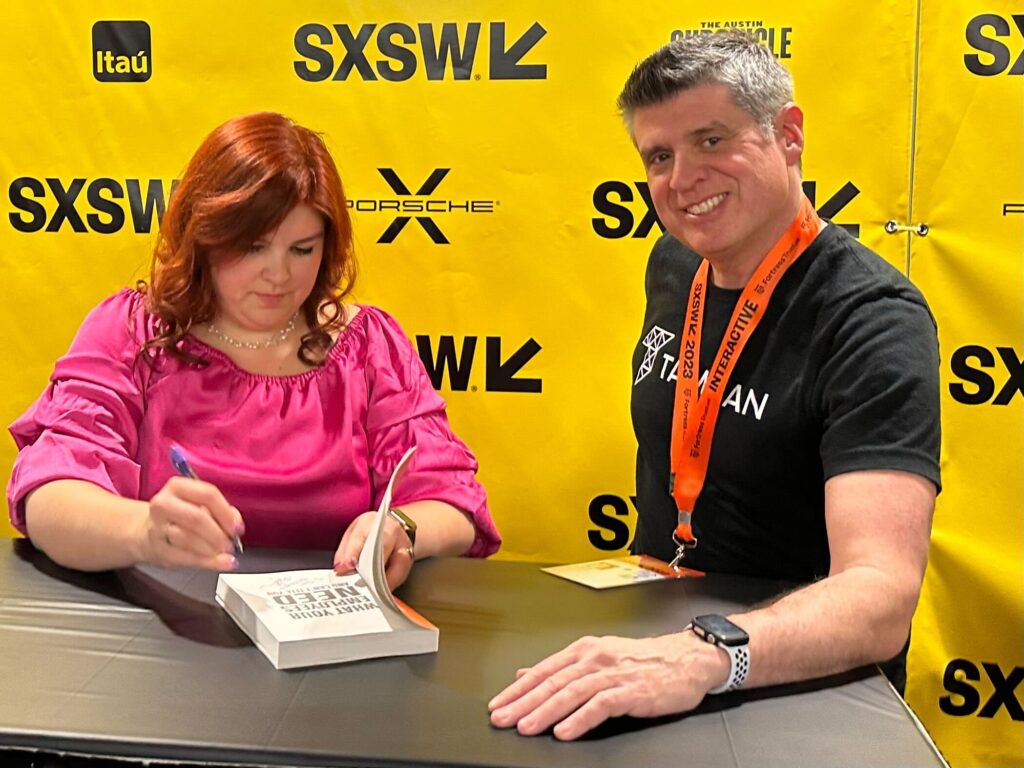 Tamman CEO, Jeff Tamburino beams towards the camera while wearing a Tamman shirt. Standing next to him is author Melina Palmer, who is signing a copy of her book, What Your Employees Need and Cannot Tell You, for Jeff.