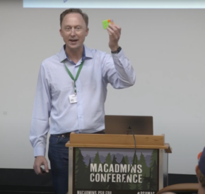 Tobias Morrison speaks on his experience as a Senior Mac Enterprise Engineer Lead at MacAdmins 2023 Conference. He is dressed in a blue button-down shirt, navy slacks and is holding a completed Rubix cube.