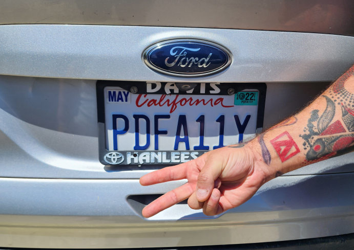 Accessible Document Specialist Dax Castro illustrates his passion for accessibility as makes a peace sign in front of his Ford car's license plate, which reads "PDFA11Y". His arm has a sleeve of tattoos, and he is showing his Adobe Suite logo tattoo prominently.