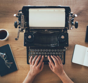 A writer uses a well-maintained typewriter. Next to them are glasses, black coffee, an open notebook with a pencil, and a smartphone.