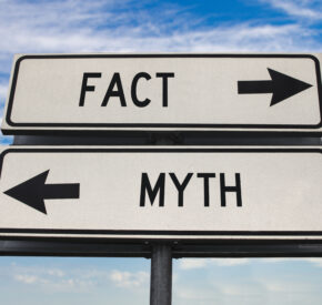 Two rectangular white street signs on top of each other. The top sign reads "Fact" with a right facing arrow in black writing. Below it reads "Myth" with a left facing arrow, also in black writing.