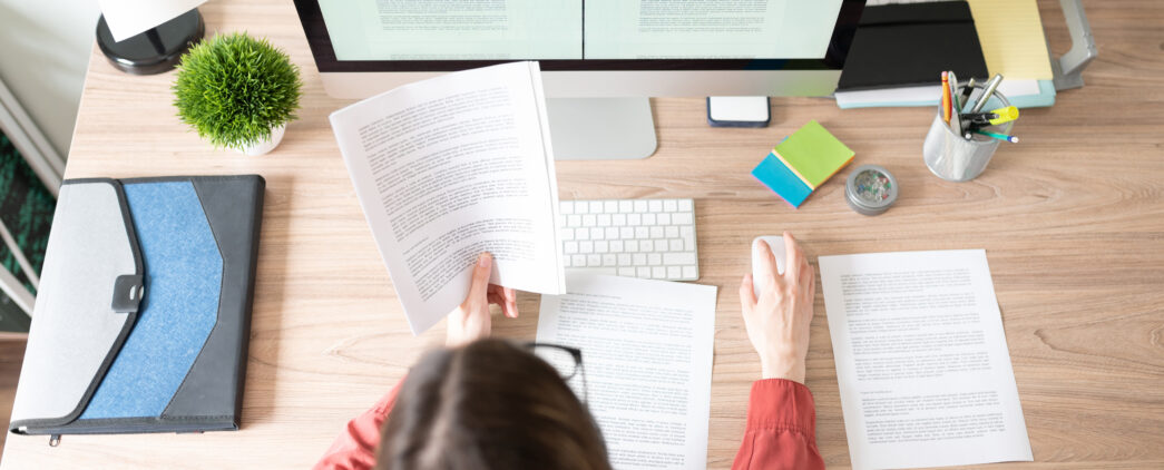 A digital document remediation subject matter expert changes a document to be more accessible to someone at her desk. In her hand is a printed copy of the document she is remediating on her computer.