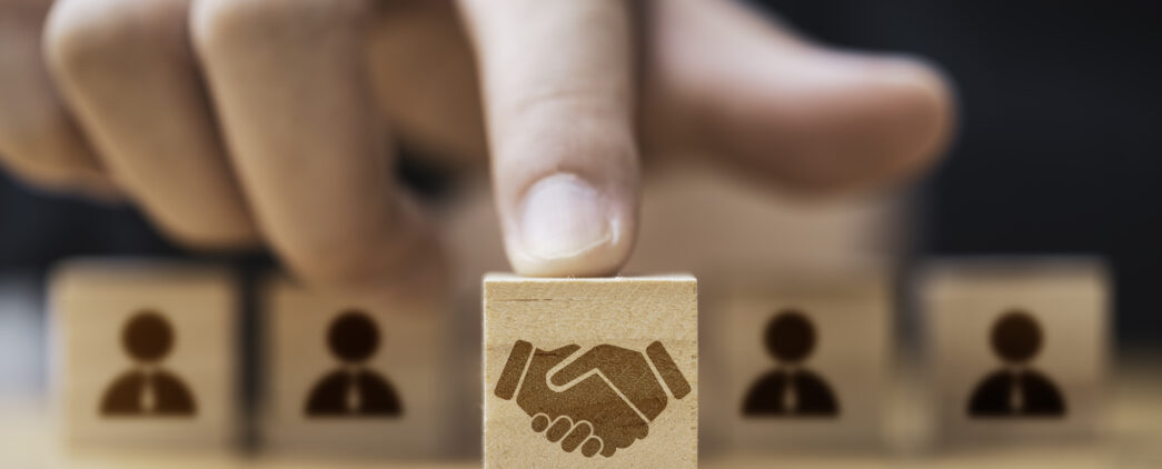 A corporate decision maker uses their pointer finger to push a small wooden cube forward. The cube is 3rd in a series of 5 cubes, and has a brown image of people shaking hands on it. The other 4 blocks are in the background and depict people in ties.