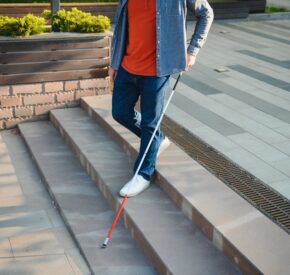 In a city, a blind traveler independently navigates an outdoor plaza, using their cane as they walk down some stairs.