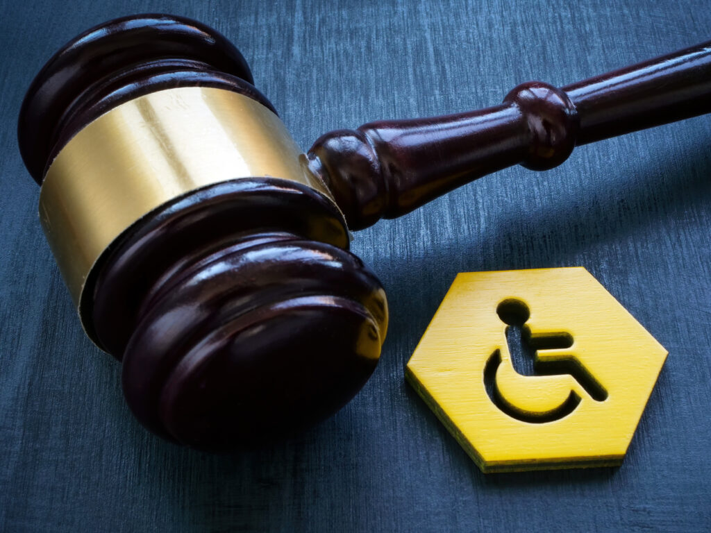 A brown legal gavel with a gold stripe along the middle where the hammer is. To the lower right of the gavel is a gold colored, wooden hexagon with the symbol of a person in a wheelchair hollowed into the hexagon.