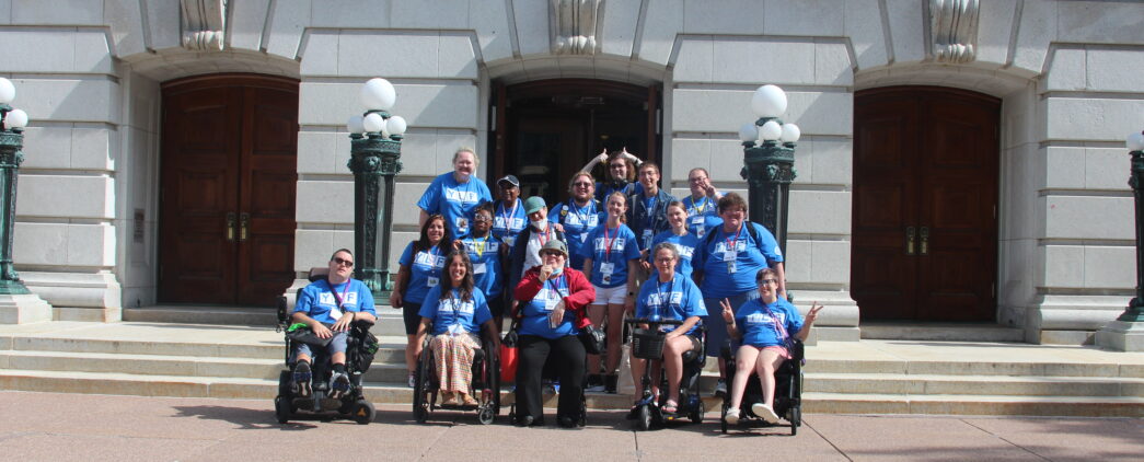 A group of people, including a number in wheelchairs, from the Wisconsin Youth Leadership Forum, wearing matching blue shirts smile in front of the Capitol Building in Madison.