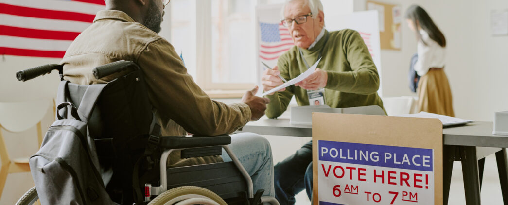 A man in a wheelchair is talking to a poll worker seated at a table inside a polling place. An American flag is displayed on the wall, and a sign on the table reads, 'Polling Place, Vote Here! 6 AM to 7 PM Today.'