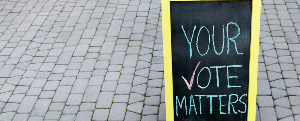 An A-frame chalkboard sign reads 'YOUR VOTE MATTERS,' with a checkmark next to the word 'VOTE. The yellow-framed sign is situated on a paved walkway.