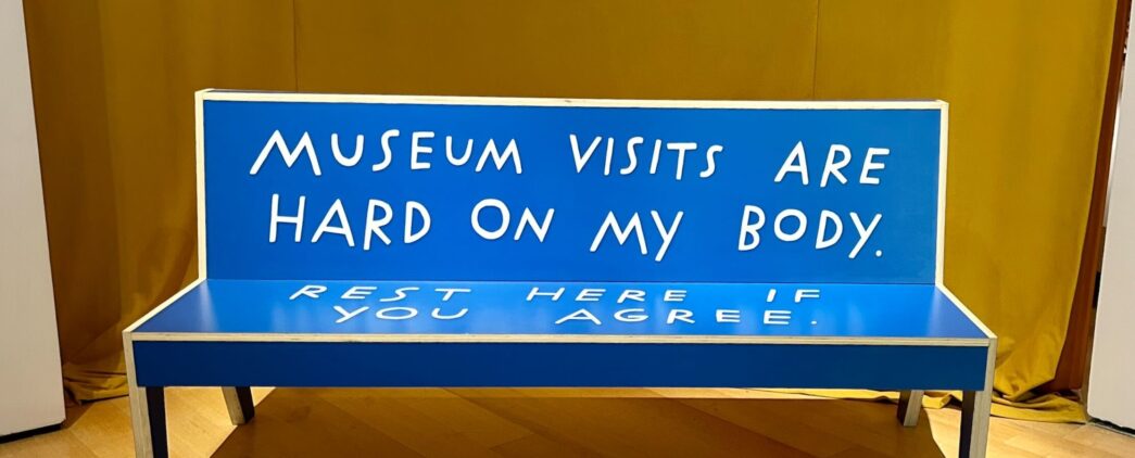 A six foot long wooden bench with straight edges colored blue with large capital letters that read: Museum visits are hard on my body rest here if you agree.