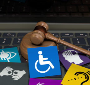 A wooden gavel resting on a laptop keyboard surrounded by colorful accessibility icons, including symbols for wheelchair access, sign language, visual and hearing impairments, and elderly care.