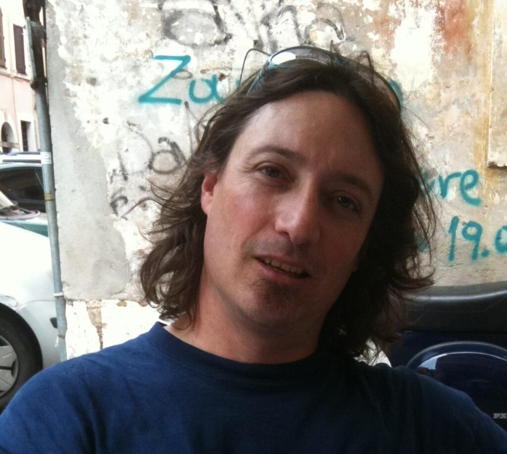 Markus Goldman half smiling at the camera in a blue shirt with some graffiti on the wall behind him