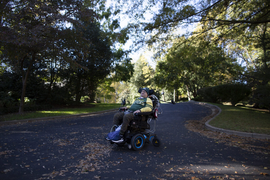 Jim Isenberg wearing a Packers shirt and beanie while sitting in his electric wheelchair in a long tree-lined driveway in the fall.